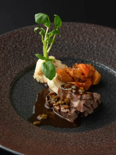 黒色の皿に盛られた、肉料理。焼き色がついた肉の上にソースがかかっており、マッシュポテトと野菜のソテーが添えられている。