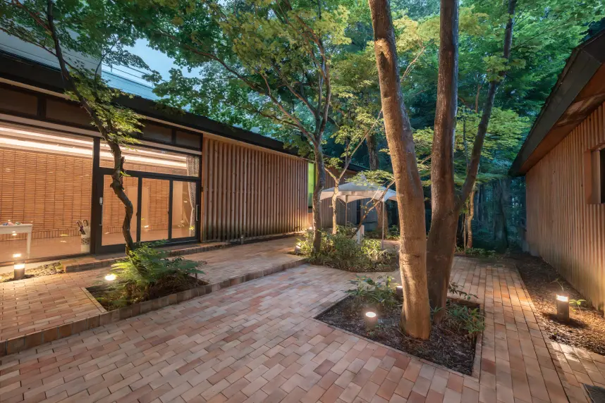 緑豊かな木々に囲まれた旅館の中庭。レンガ敷きの小道があり、テーブルと椅子が並ぶ開放的な空間。